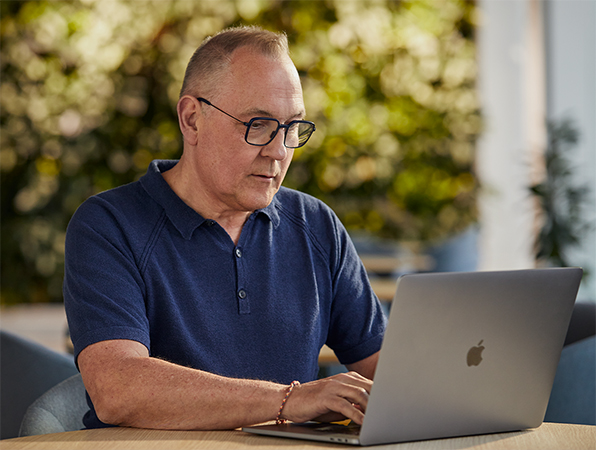 Funcionário da equipe de software e serviços da Apple trabalha em um MacBook na área externa com folhagens ao fundo.