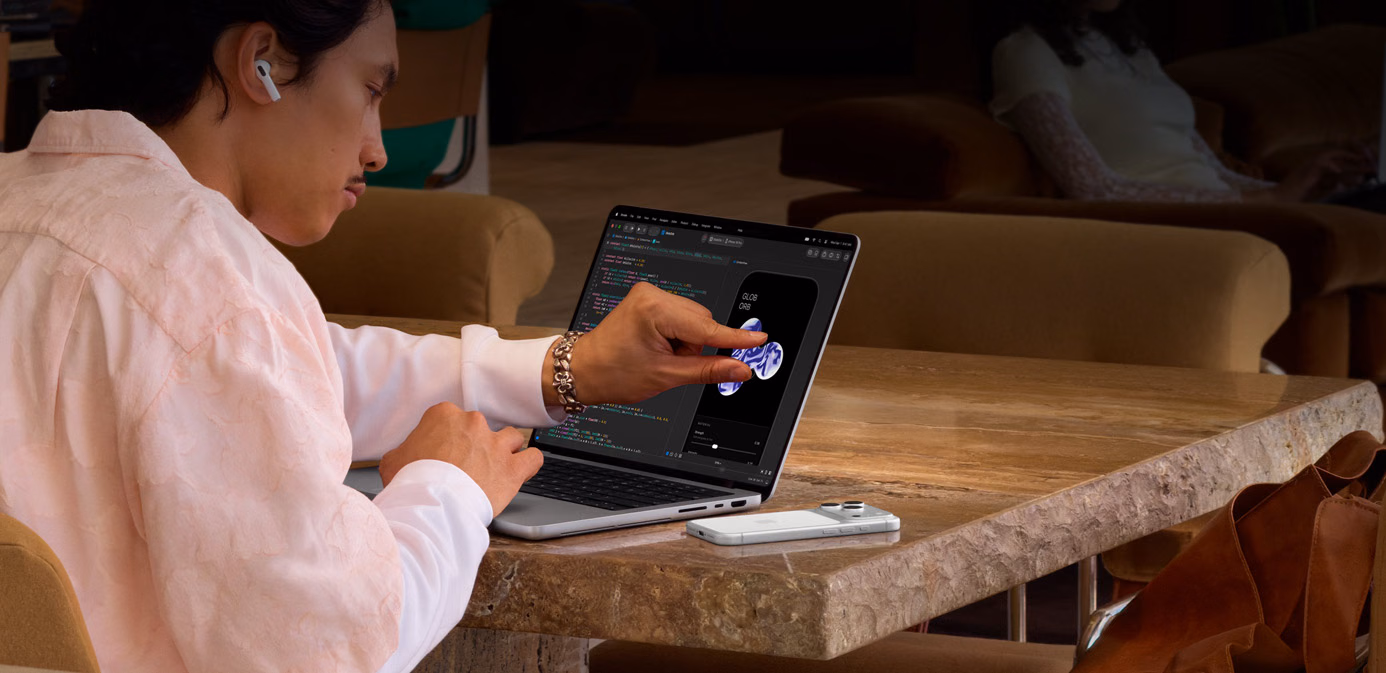 Henkilö istuu pöydän ääressä julkisessa tilassa ja säätää nipistyseleen avulla Xcoden elementtiä käyttämällä hopeanvärisen PROLED XDR  ‑näytöllä varustetun MacBook Pron kosketusominaisuutta, pöydällä kannettavan vieressä on iPhone, ja henkilö käyttää AirPods Pro ‑kuulokkeita