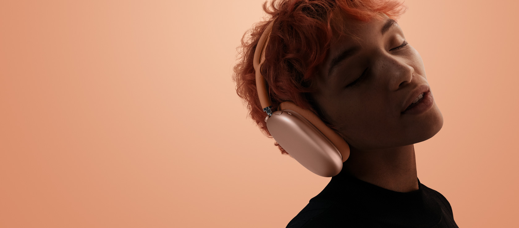 O persoană cu capul înclinat și ochii închiși poartă AirPods Max 2 pe Portocaliu, pe un fundal portocaliu-roz