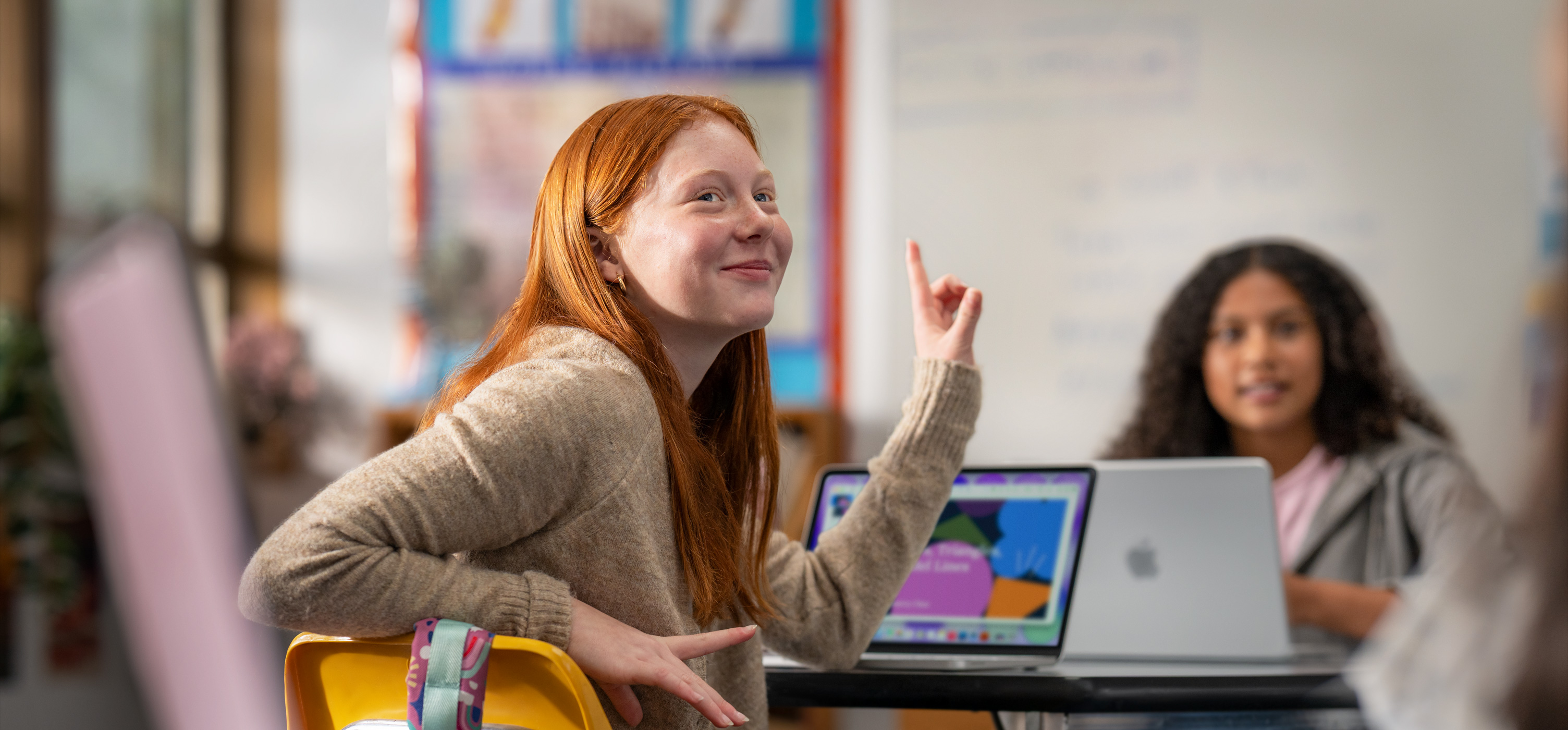 Una estudiante sentada frente a un Macbook en un salón de clases levanta la mano para responder una pregunta, se muestra la facilidad de uso y la participación en clase