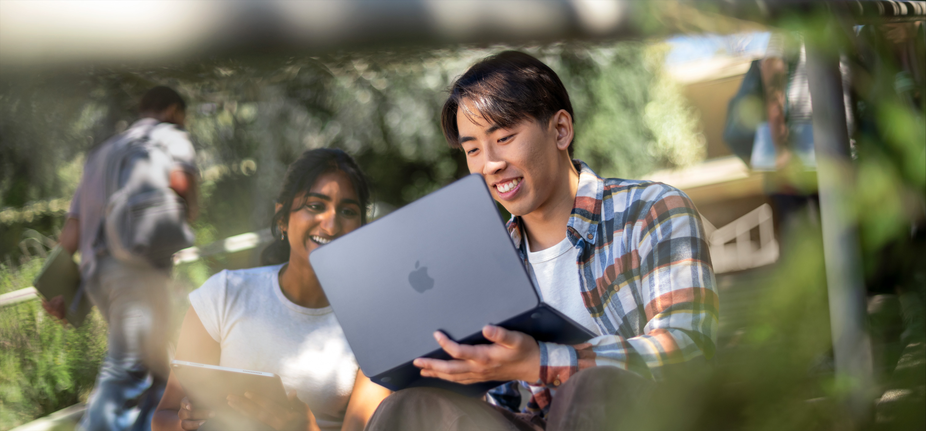 Dos estudiantes sonríen mientras usan un MacBook al aire libre, uno la sostiene en su regazo mientras colaboran en un proyecto
