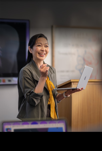 Una profesora hace una presentación a sus estudiantes en un salón mientras sostiene un Macbook color plata, se muestra el rendimiento y la larga duración de la batería