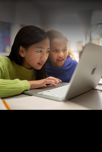 Dos estudiantes de primaria en un escritorio miran con atención un Macbook mientras trabajan en un salón de clases