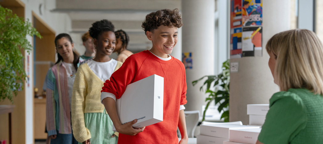 Un estudiante recibe un MacBook entregado por el equipo de sistemas, otros estudiantes hacen fila detrás de él, se destaca la facilidad de implementación