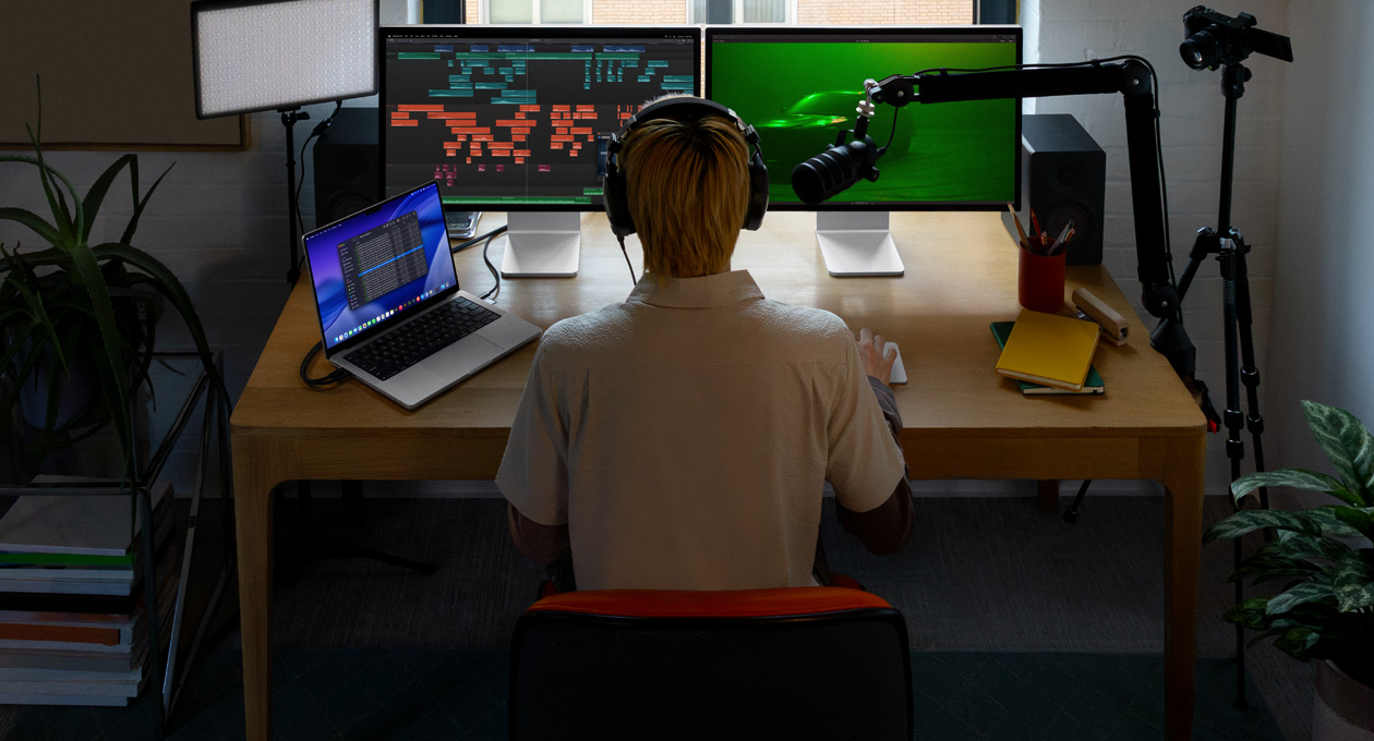 Eine Person sitzt an einem Schreibtisch mit einem Mikrofon und Kopfhörern vor drei Geräten, einem Mac Studio Display Bildschirm, Tools zum Bearbeiten von Audio, Soundclips in verschiedenen Farbblöcken, einem zweiten Mac Studio Display Bildschirm, der eine stimmungsvolle Aufnahme eines Autos im Nebel mit grünem Licht zeigt, einem 16 Zoll MacBook Pro Bildschirm, der die Finder App auf dem Schreibtisch mit Dateien für Final Cut Pro zeigt, Dock, stilisierter blauer Schreibtischhintergrund