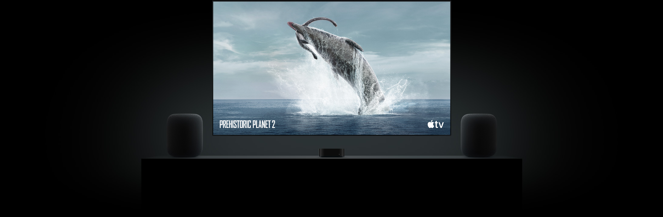 Large flat-screen TV featuring a vivid image of a dinosaur from Prehistoric Planet. The TV is hung above an Apple TV 4K and framed by two HomePod speakers placed on a living room console