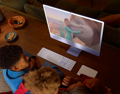 Three young people sitting at a table with an iMac, Magic Keyboard, and Magic Trackpad, watching a movie